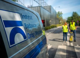 Zbliżenie na logo IBDiM na samochodzie służbowym, w tle pracownicy wykonujący badania stanu oznakowania poziomego na drodze.