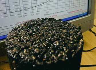 Kostka kruszywa asfaltowego, a w tle monitor komputera wyświetlający wyniki pomiarów badawczych.