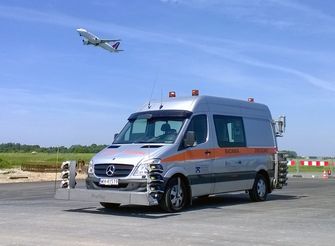 Samochód dostawczy do badań drogowych stoi na płycie lotniska, nad nim przelatuje samolot.